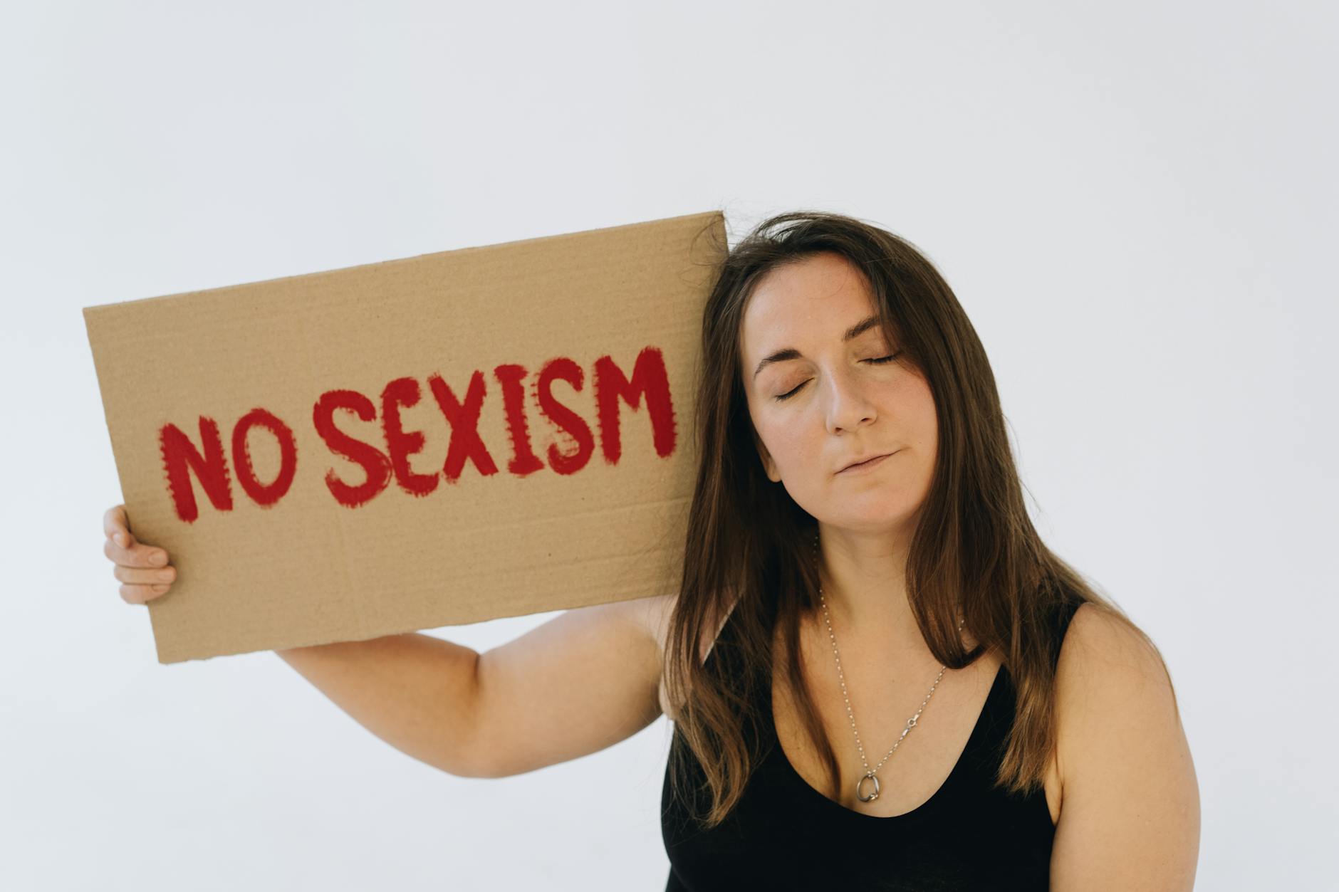 parità di genere — A woman with closed eyes holds a protest sign saying 'No Sexism' against a white background.