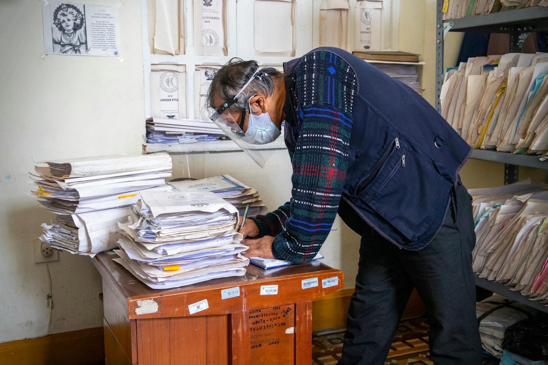 riforma pensioni 2027 — Elderly man with face shield sorts through piles of documents in an office setting.