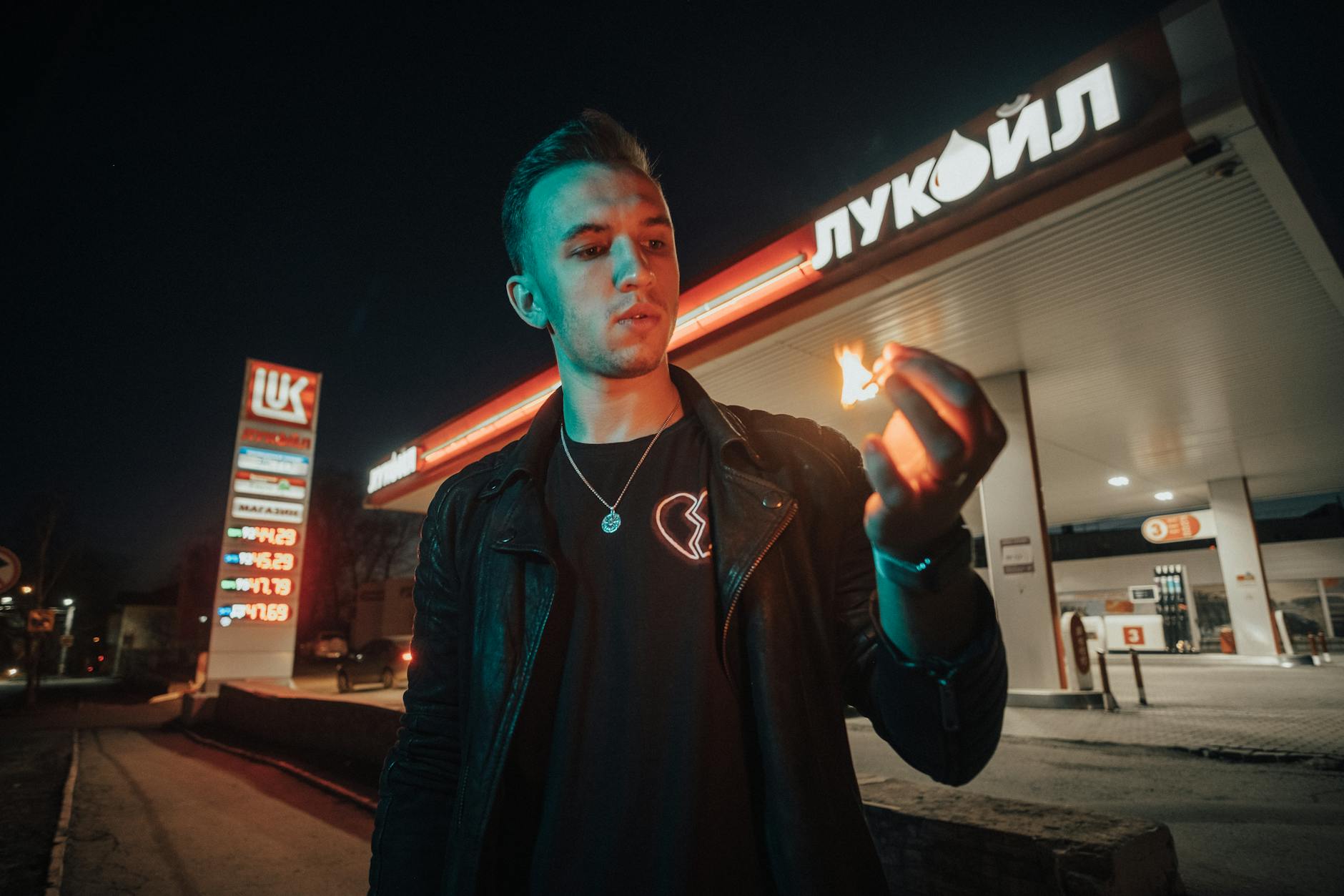 prezzi carburanti — A man holding a lit matchstick stands in front of a Lukoil gas station at night, creating a dramatic scene.