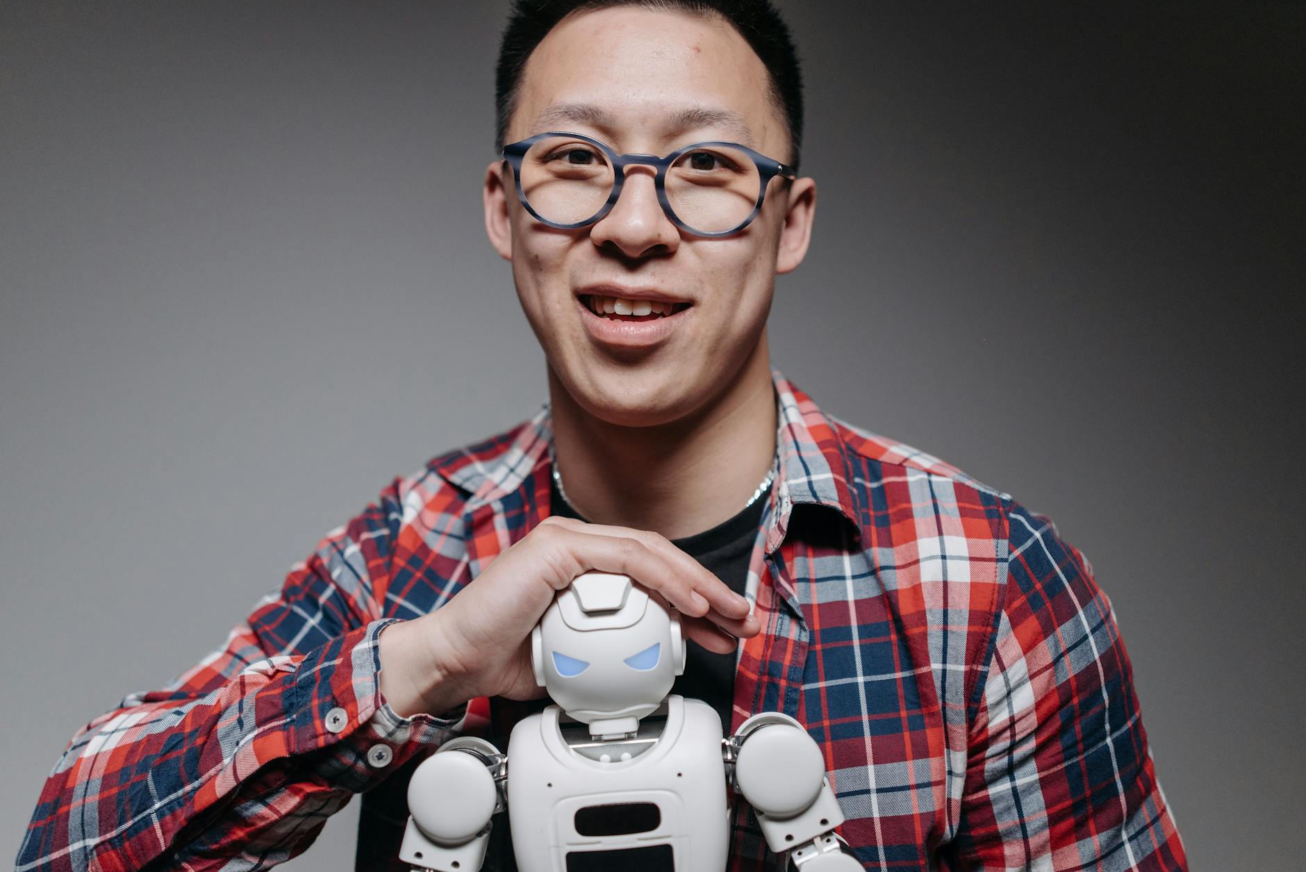 esoscheletro robotico — Asian man smiling, holding a small toy robot during a studio shoot. Indoors, with eyeglasses and plaid shirt.