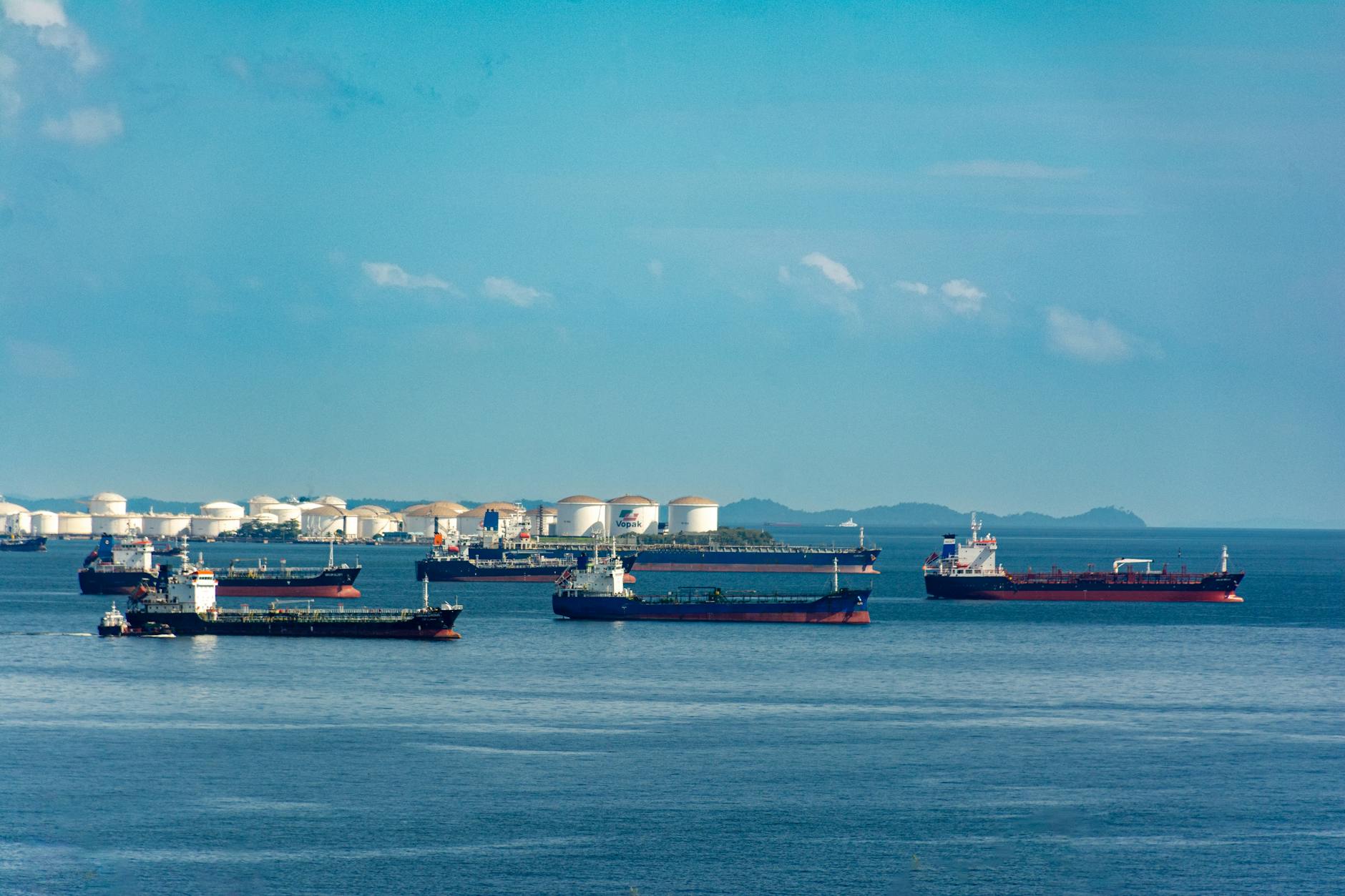 tensioni energetiche Golfo Persico — A fleet of cargo ships docked near oil storage tanks along a serene coastline with a clear blue sky above.