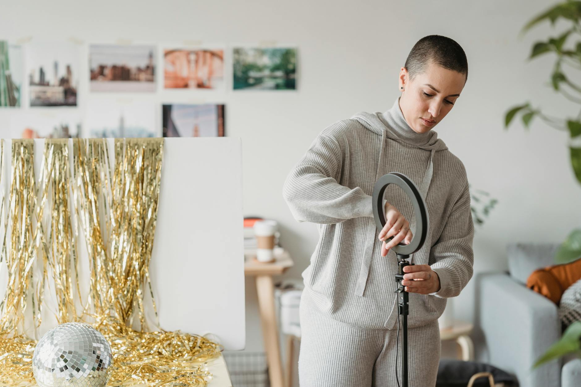ring light professionali — A woman with short hair sets up a ring light in a modern, creative home environment adorned with decorative gold tinsel.