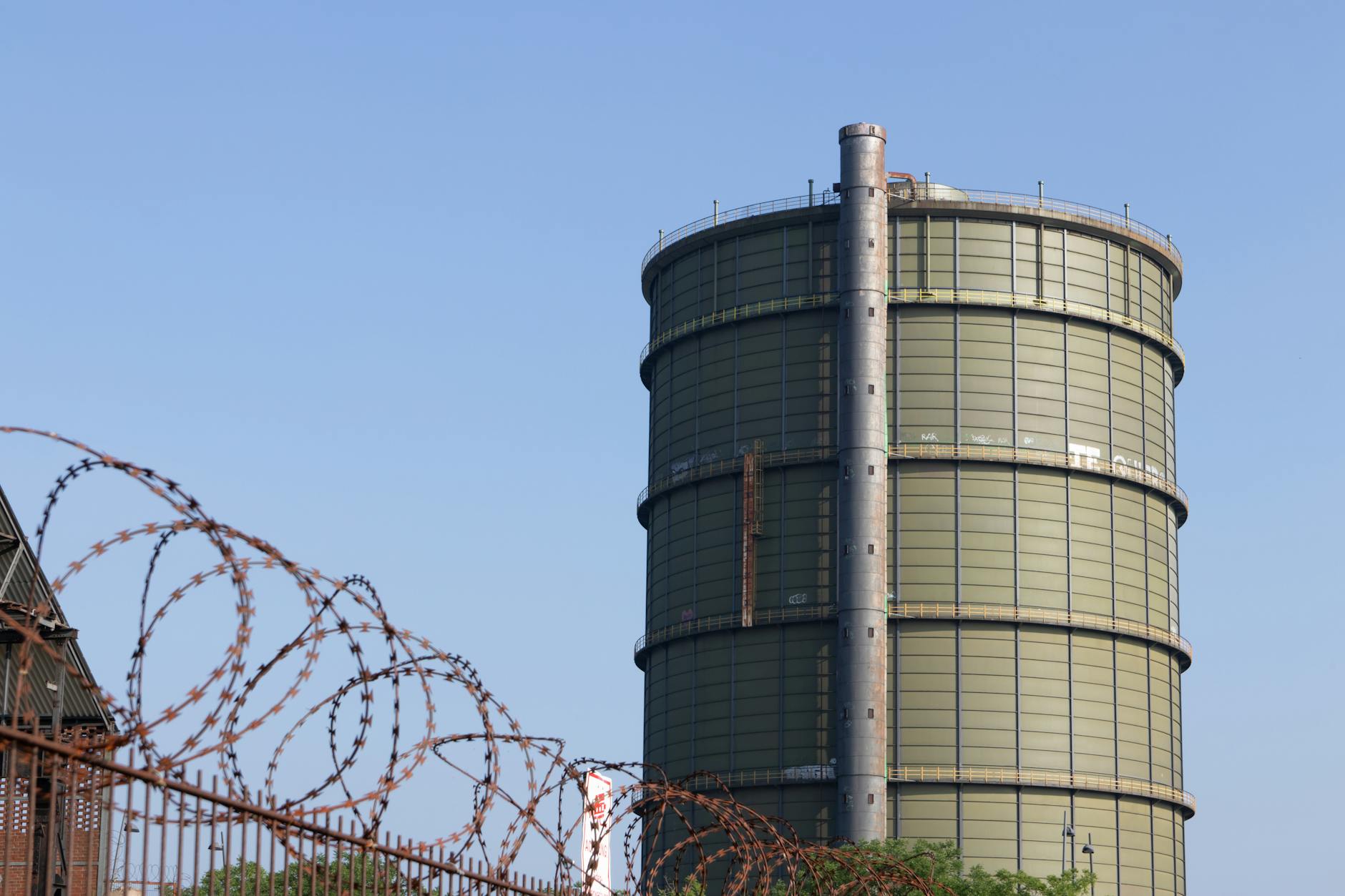 strategia europea gas — A large gas holder in an industrial area surrounded by barbed wire under a clear blue sky.