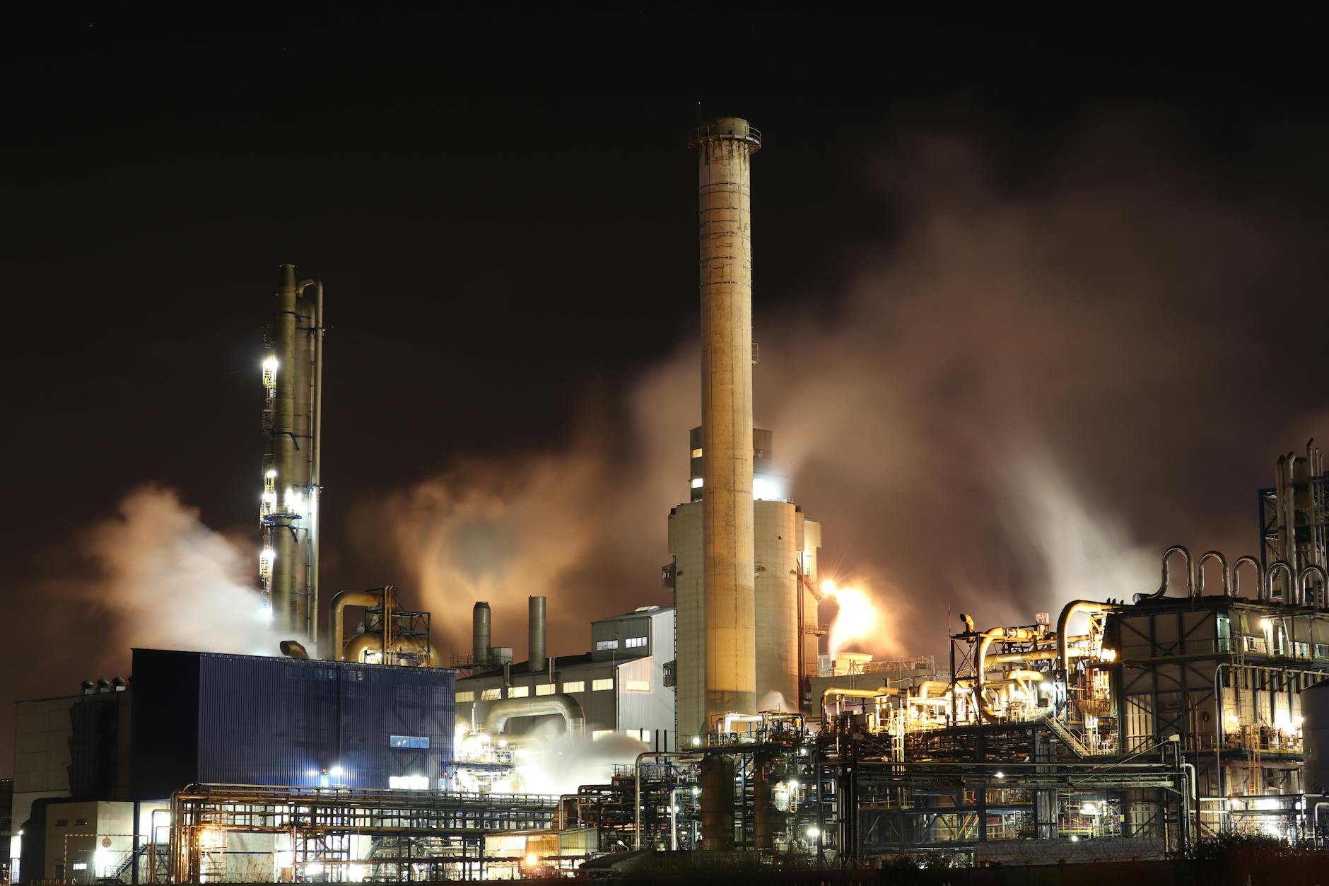 mercato energetico globale — A nighttime view of an industrial factory with smokestacks emitting smoke, highlighting energy production and pollution.