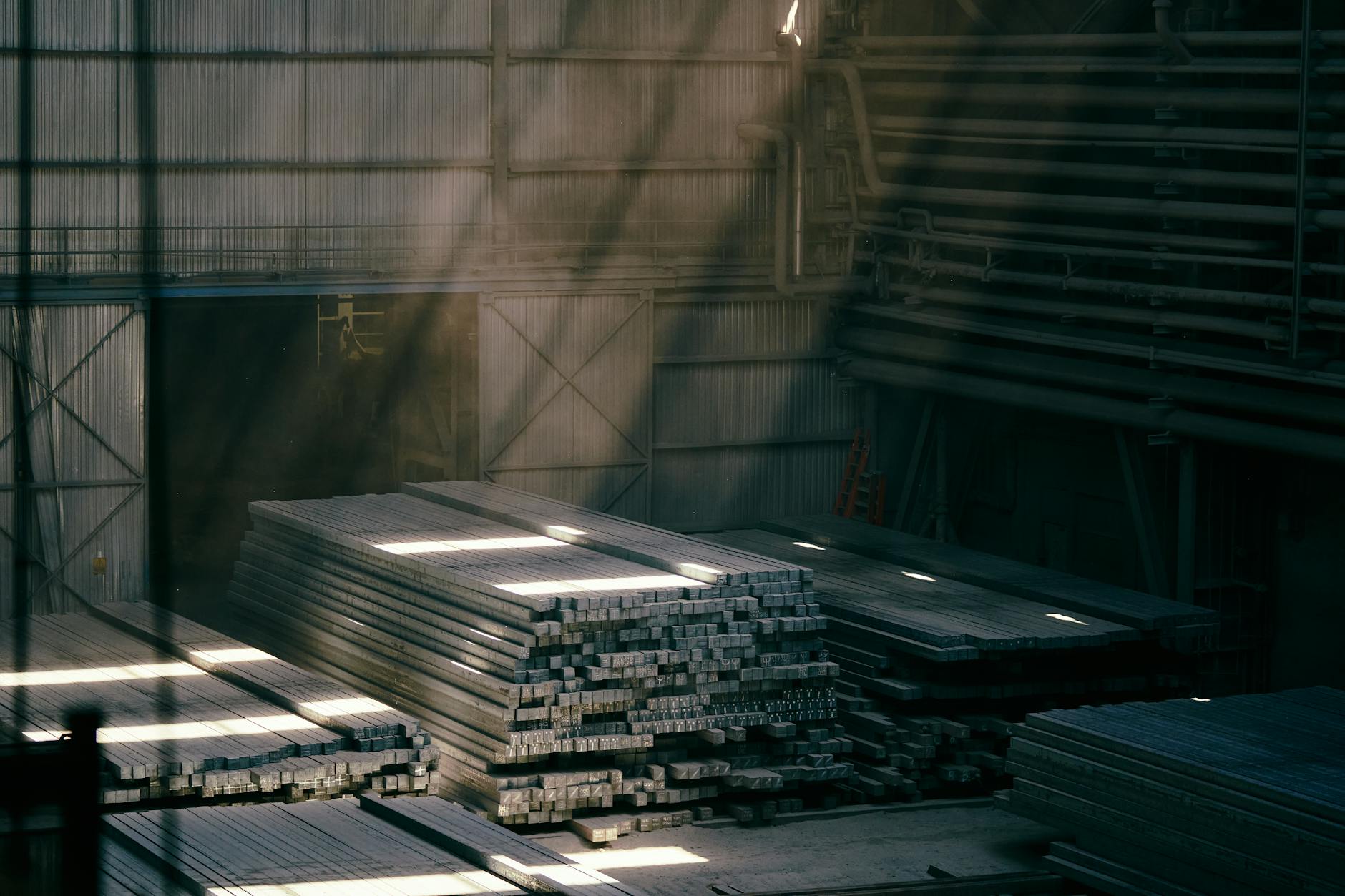 vendita Acciaierie d'Italia — Sunlight streams into an industrial warehouse illuminating stacked steel beams.