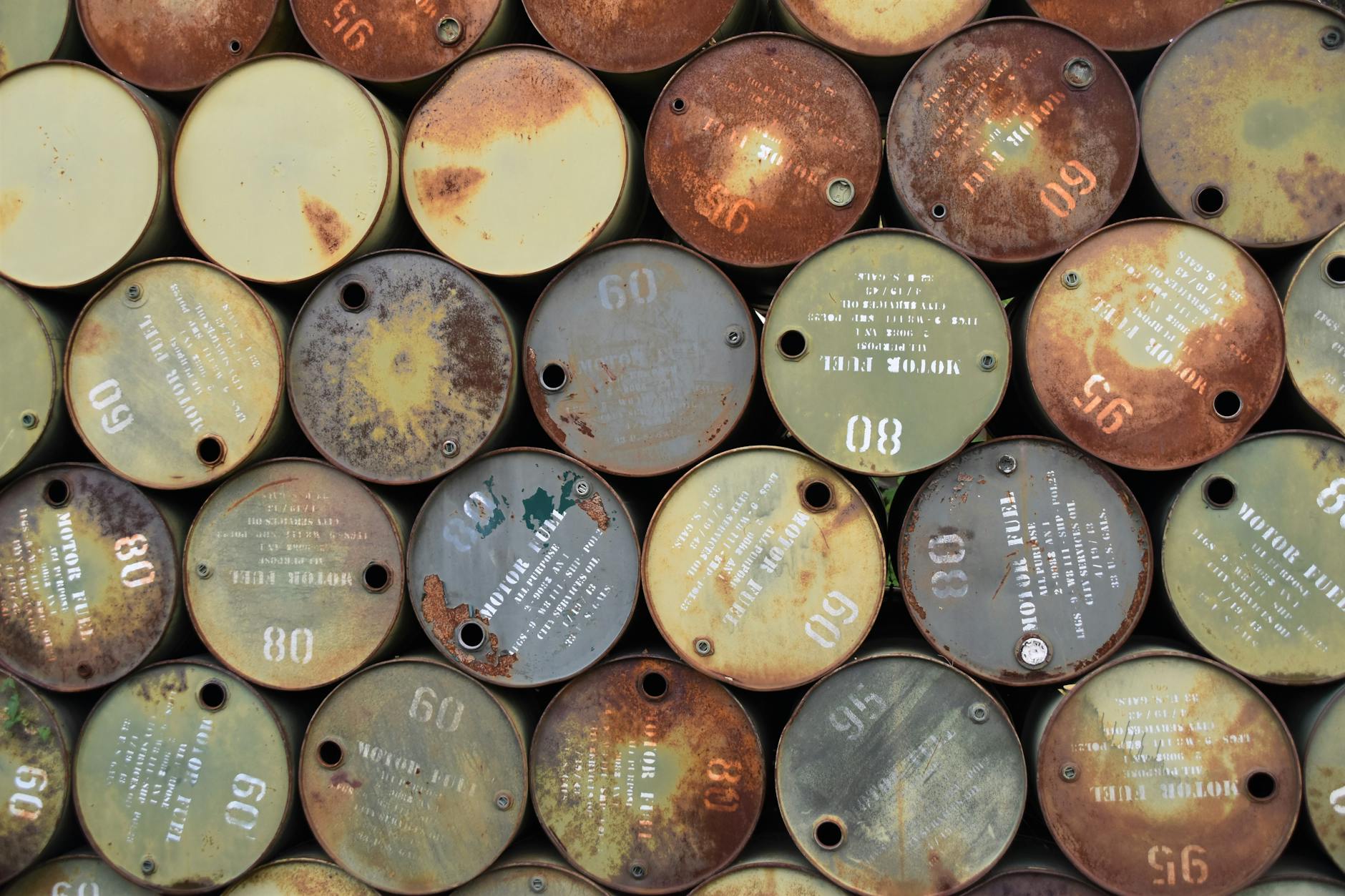 prezzi crude WTI — Pile of old, rusty oil barrels stacked outdoors with visible labels and numbers.