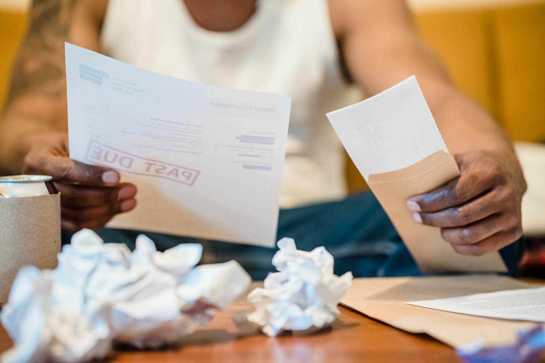 rincari energetici — Man sitting indoors reviewing past due bills with crumpled papers on a coffee table.