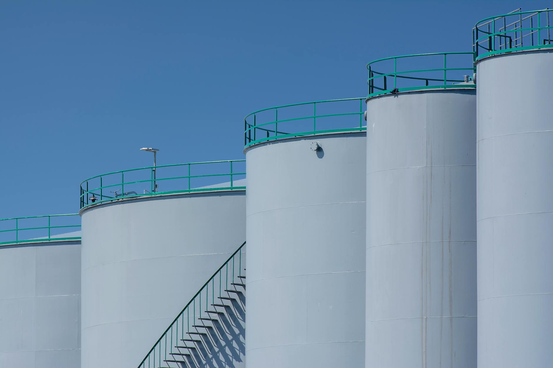 petrolio OPEC+ — Large industrial storage tanks with green railings against a clear blue sky.