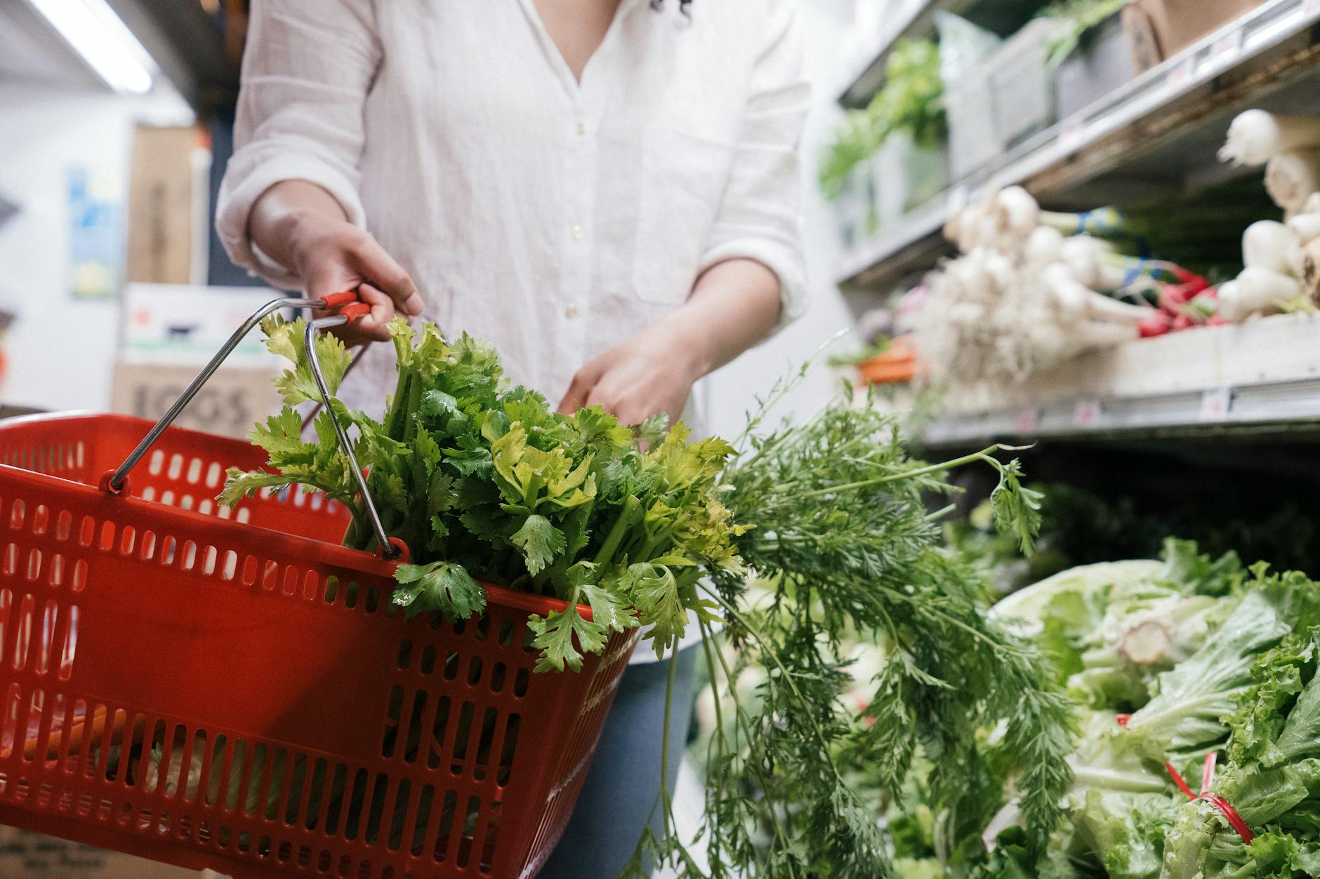 costi alimentazione sana — A person holding a grocery basket filled with fresh greens like celery and parsley in a store.