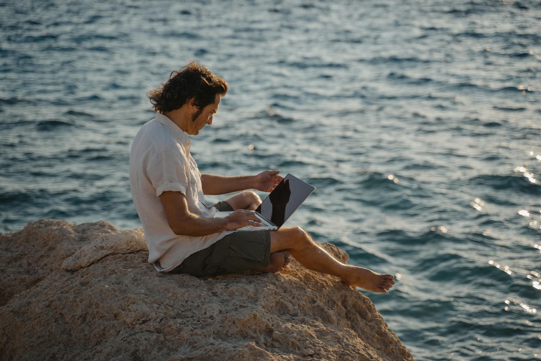 south working Sicilia — A man sitting on rocky terrain by the sea, working remotely on his laptop during the day.