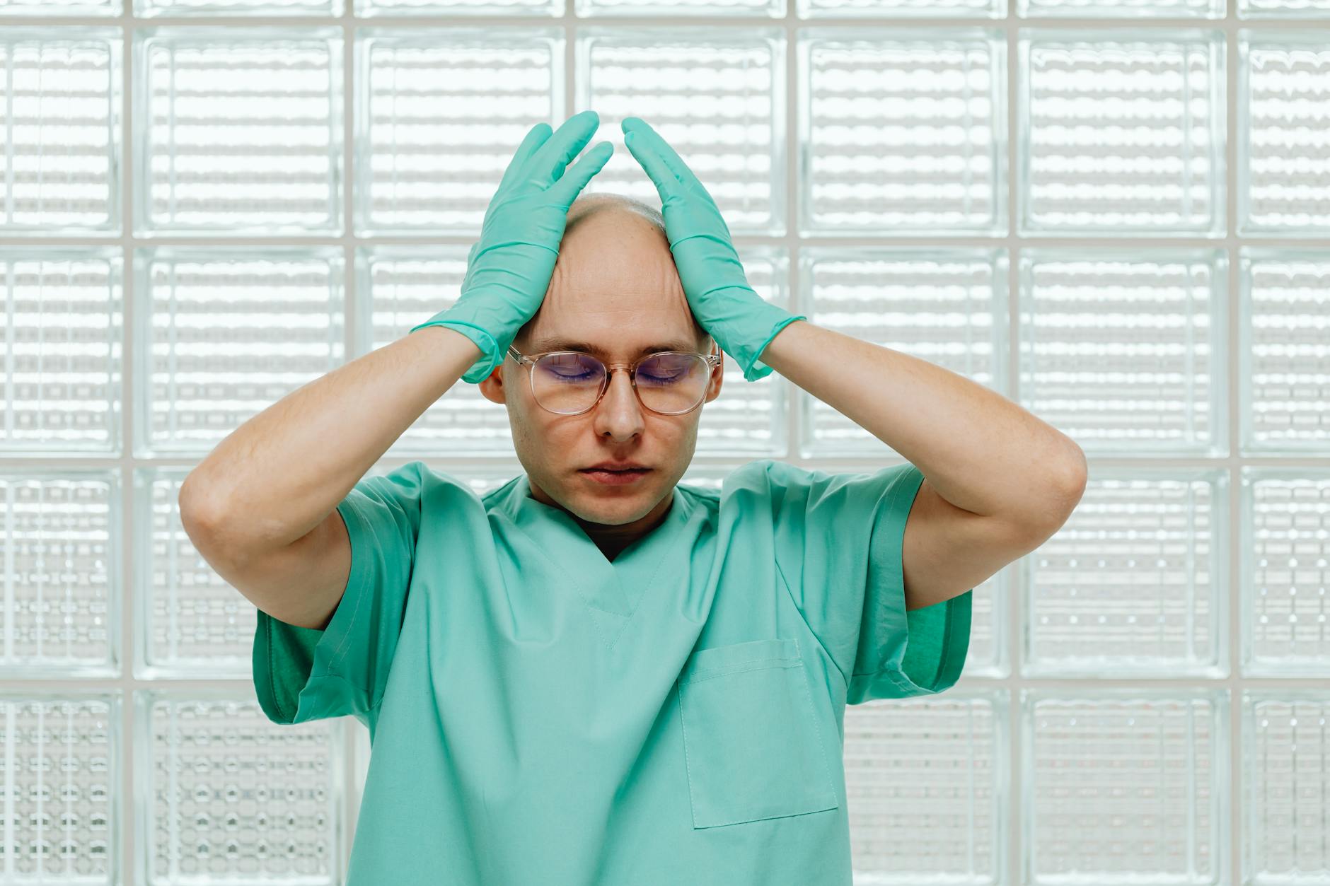 sanità italiana — A healthcare worker in green scrubs looking stressed, hands on head with eyeglasses.