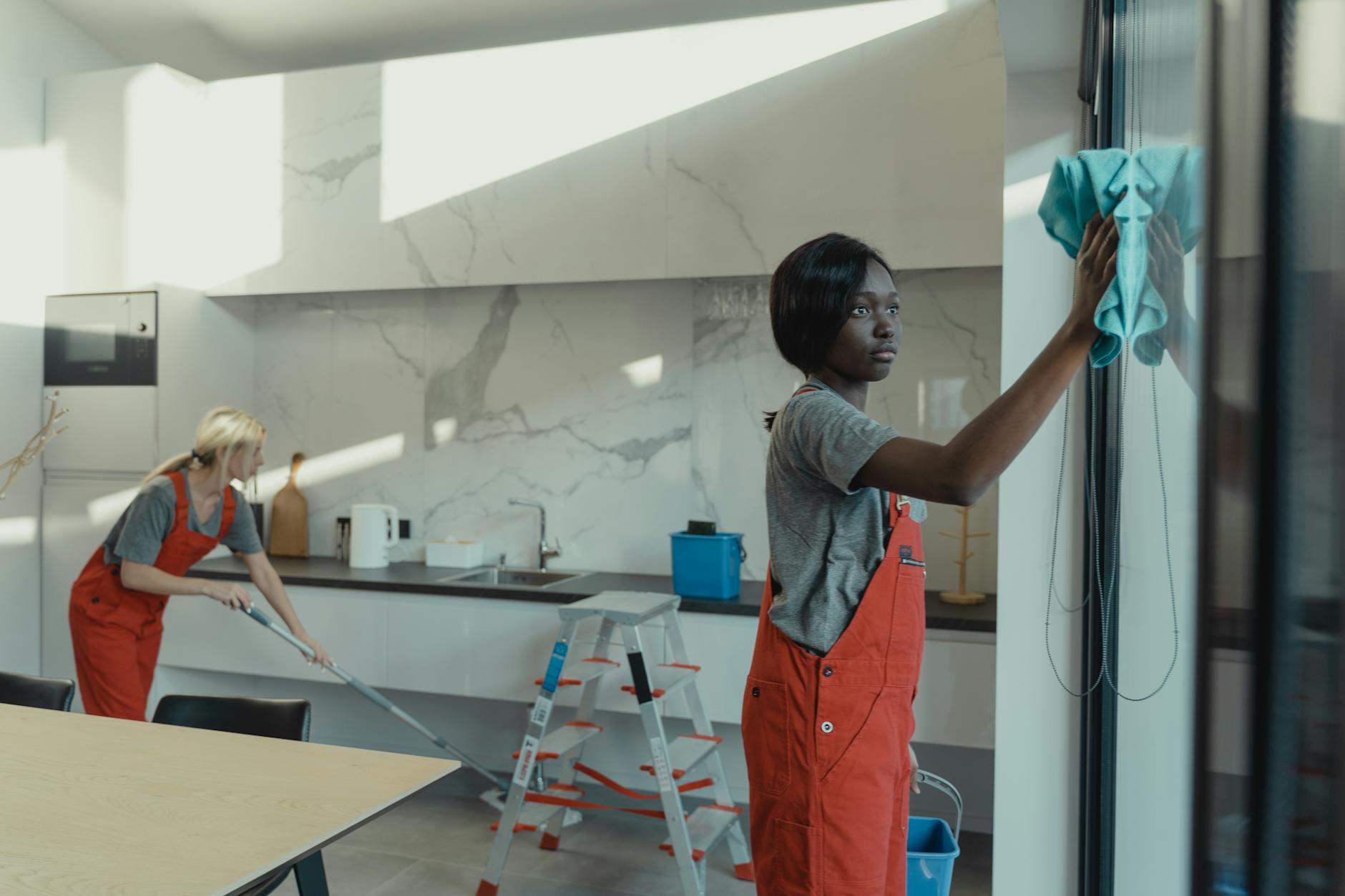 salario giusto — Two women cleaning a modern kitchen, wiping glass surfaces and mopping the floor.