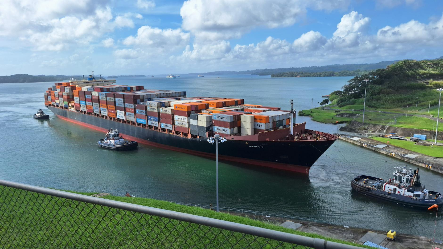 costi del canale Panama — Large container ship maneuvered by tugboats through the Panama Canal under a clear sky.