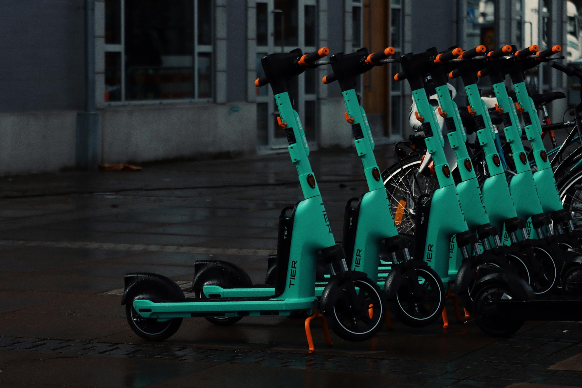 assicurazione monopattini — Multiple teal electric scooters aligned on a rainy street in Jönköping, Sweden with wet pavement.