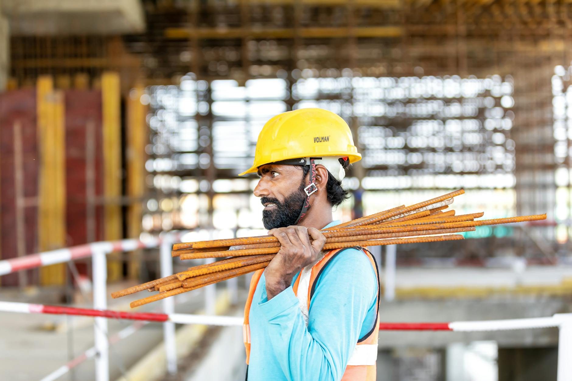sistemi IA nei cantieri — Construction worker at a building site carrying rebar, wearing a safety helmet and vest.