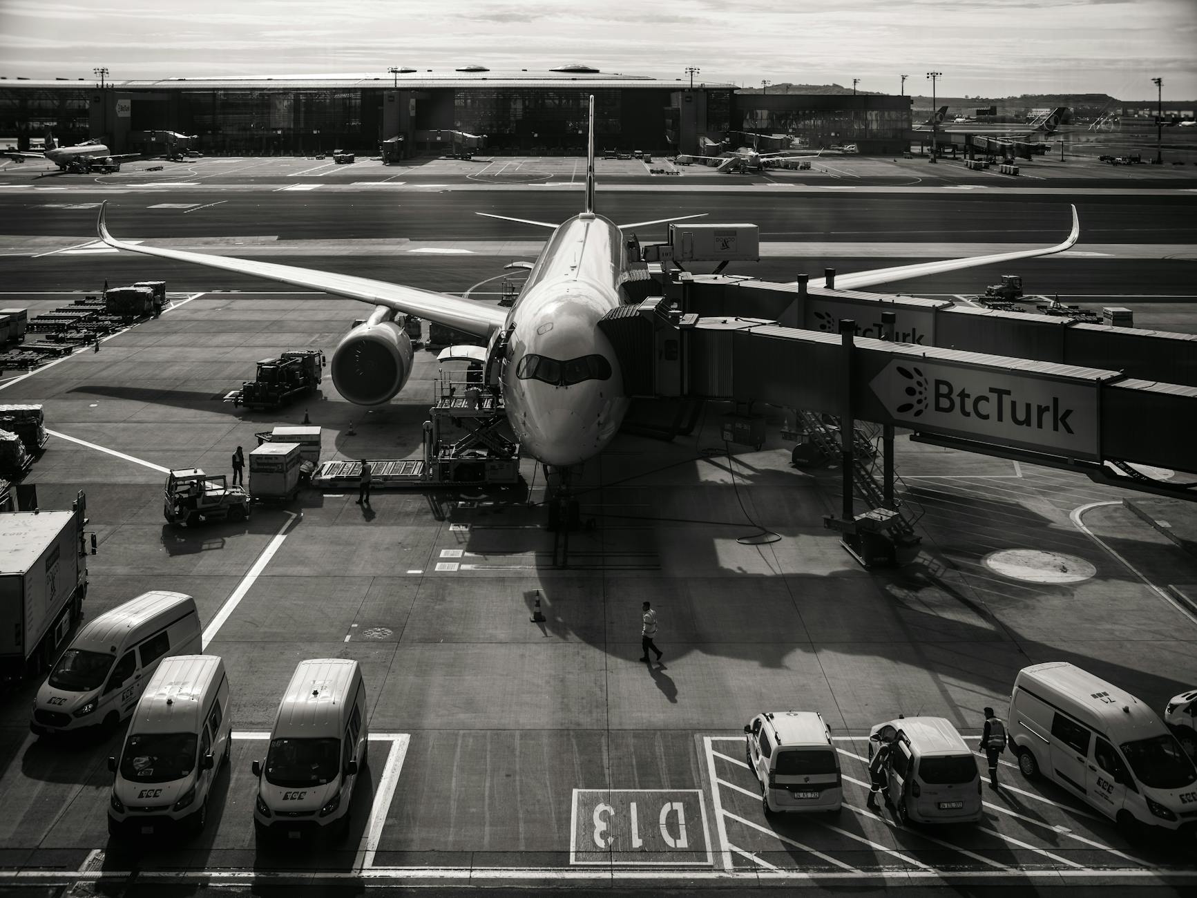 scorte carburante aviazione — Monochrome image of airplane at airport gate with vehicles and jetway during the day.
