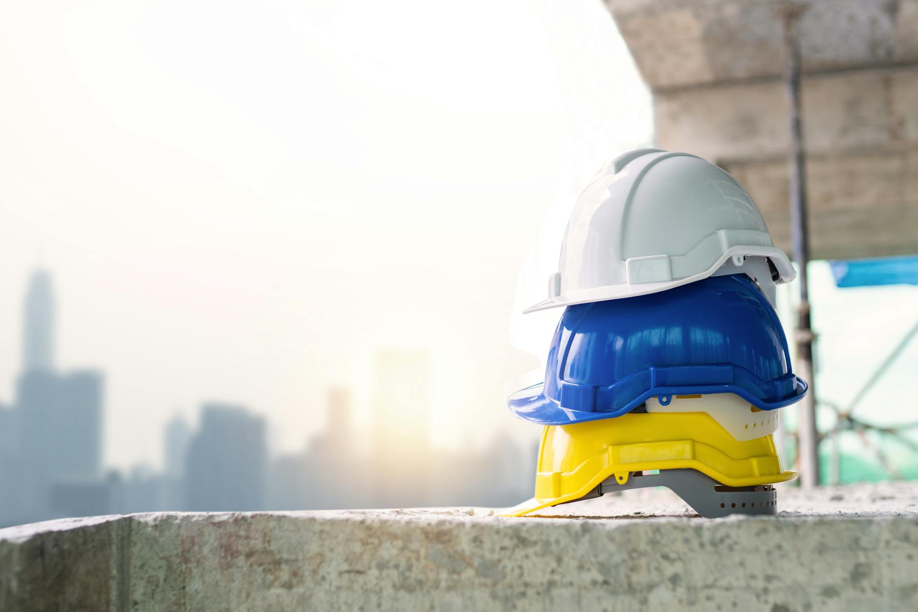 infortuni mortali sul lavoro — Three colorful safety helmets stacked on a concrete surface with city skyline in background.