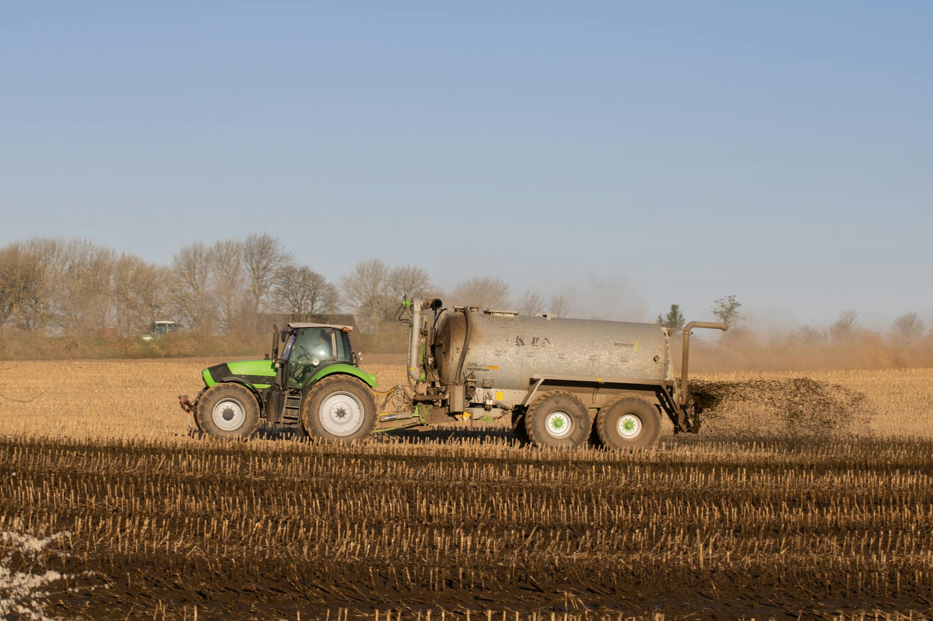 settore dei fertilizzanti agricoli — A tractor with a tanker spreading fertilizer over a barren farmland under clear skies.