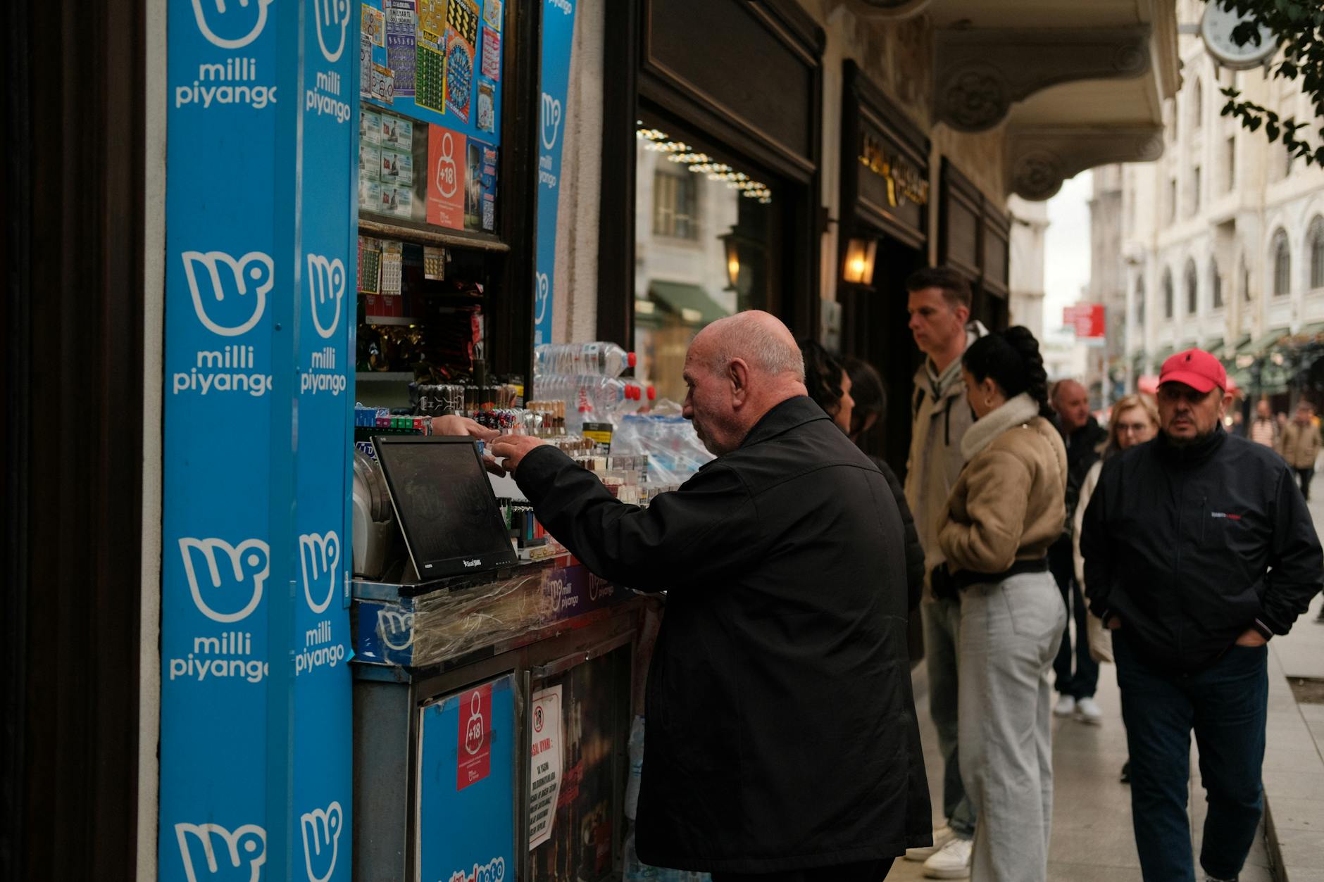 estrazione 7 maggio — People buying lottery tickets and beverages from a street vendor on a bustling urban street.