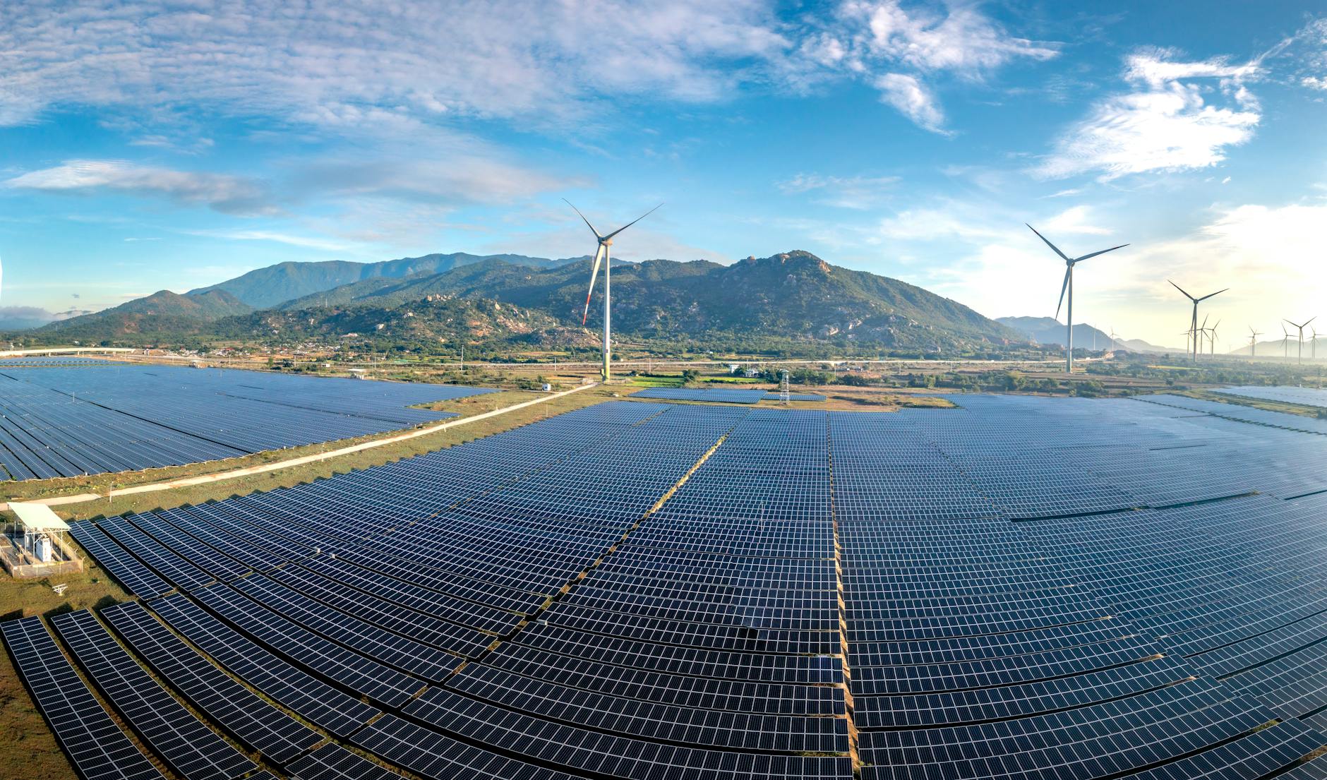 acquisizione Enel impianti fotovoltaici — Aerial view of a vast solar farm with wind turbines against a mountainous backdrop on a sunny day.