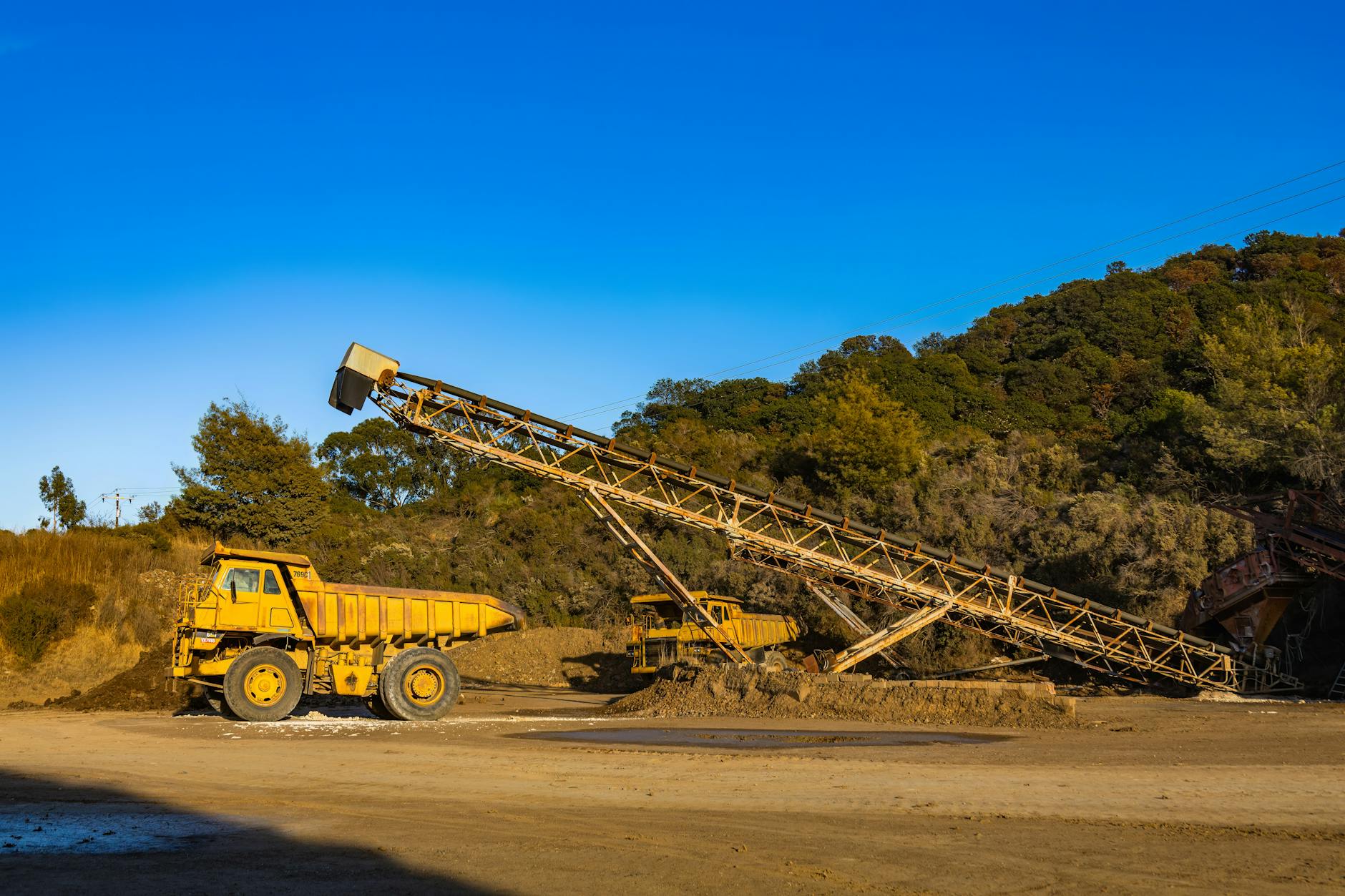 minerali critici Europa — Yellow dump trucks and conveyor machinery in a sunny mining site with clear blue sky.