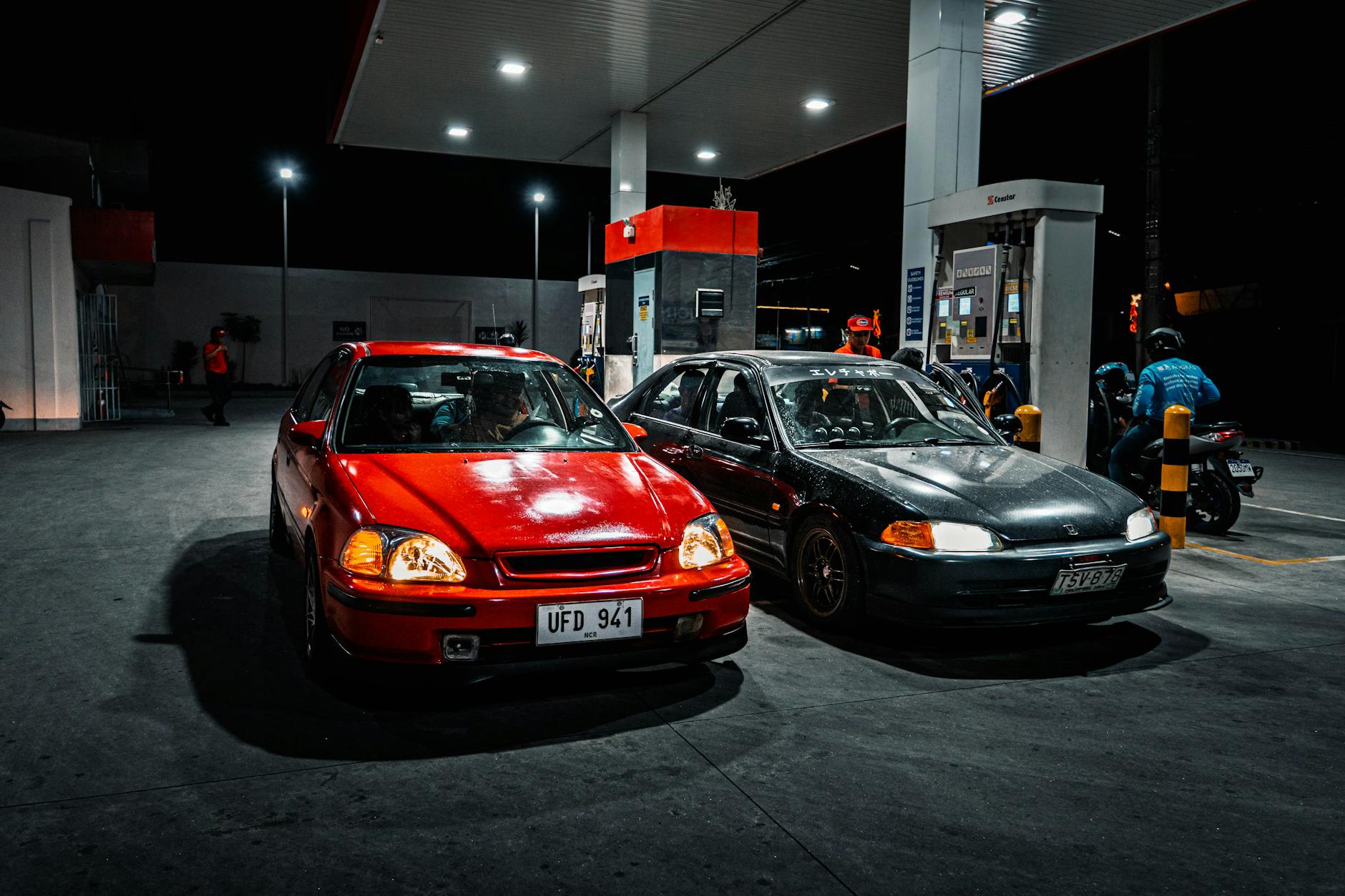 prezzi carburanti — Two cars parked at a gas station at night, illuminated by overhead lights.