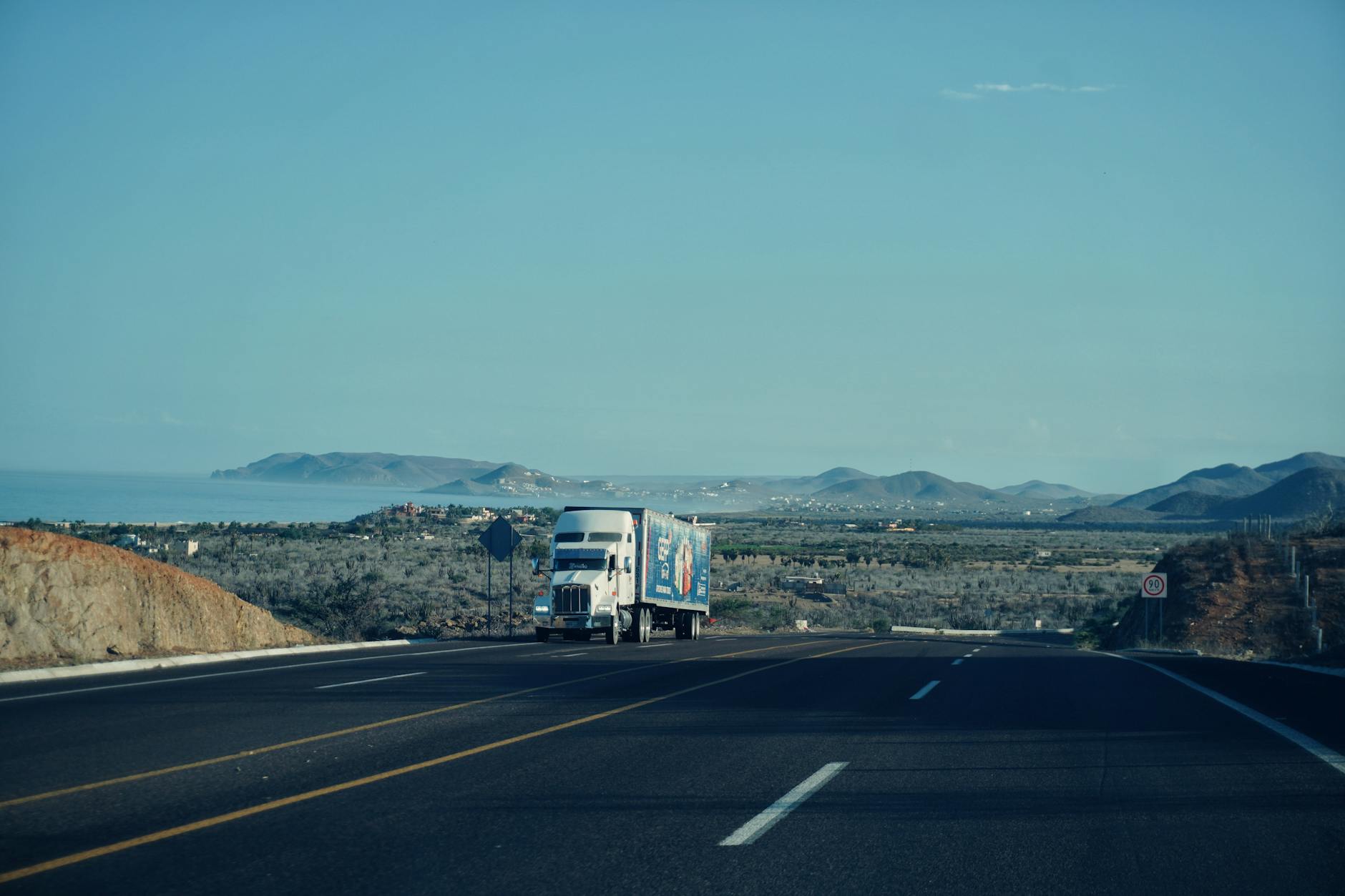 taglio delle accise — A semi-truck travels on a scenic highway with mountains in the background under a clear sky.