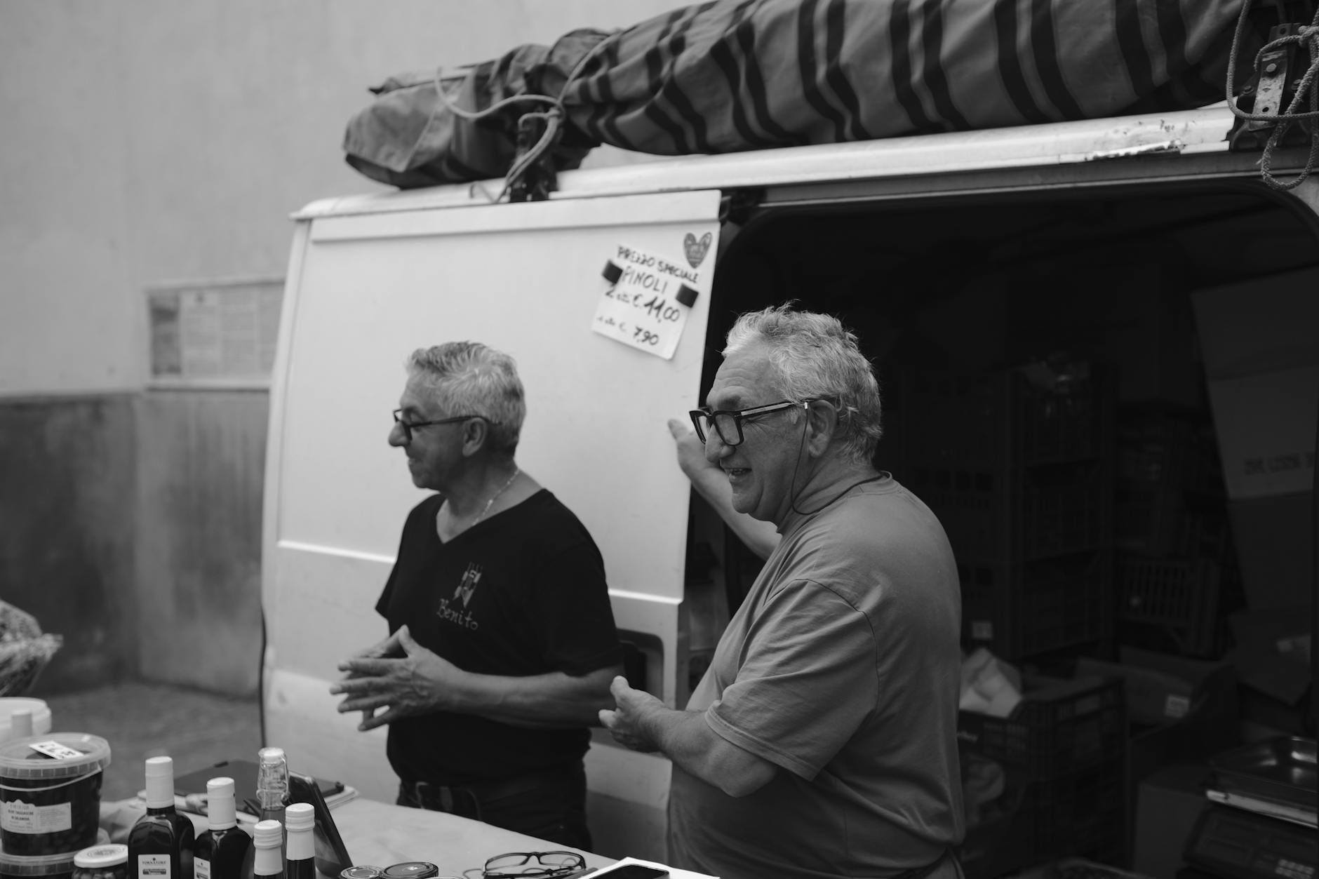 prodotti nazionali — Two vendors engaging at an Italian market stall, captured in black and white.