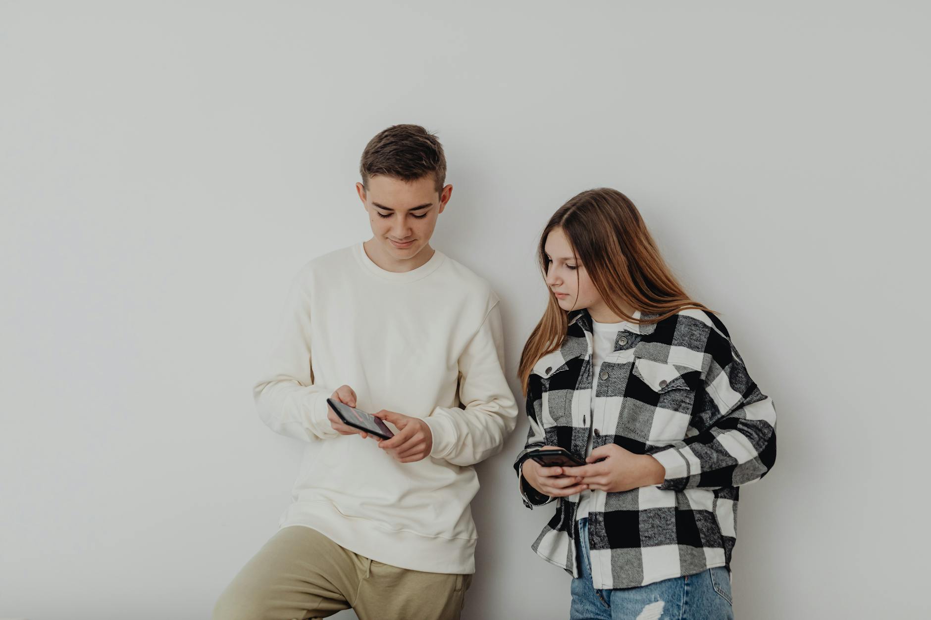 vietare social ai minori — Teenage boy and girl using smartphones while leaning against a white wall indoors.