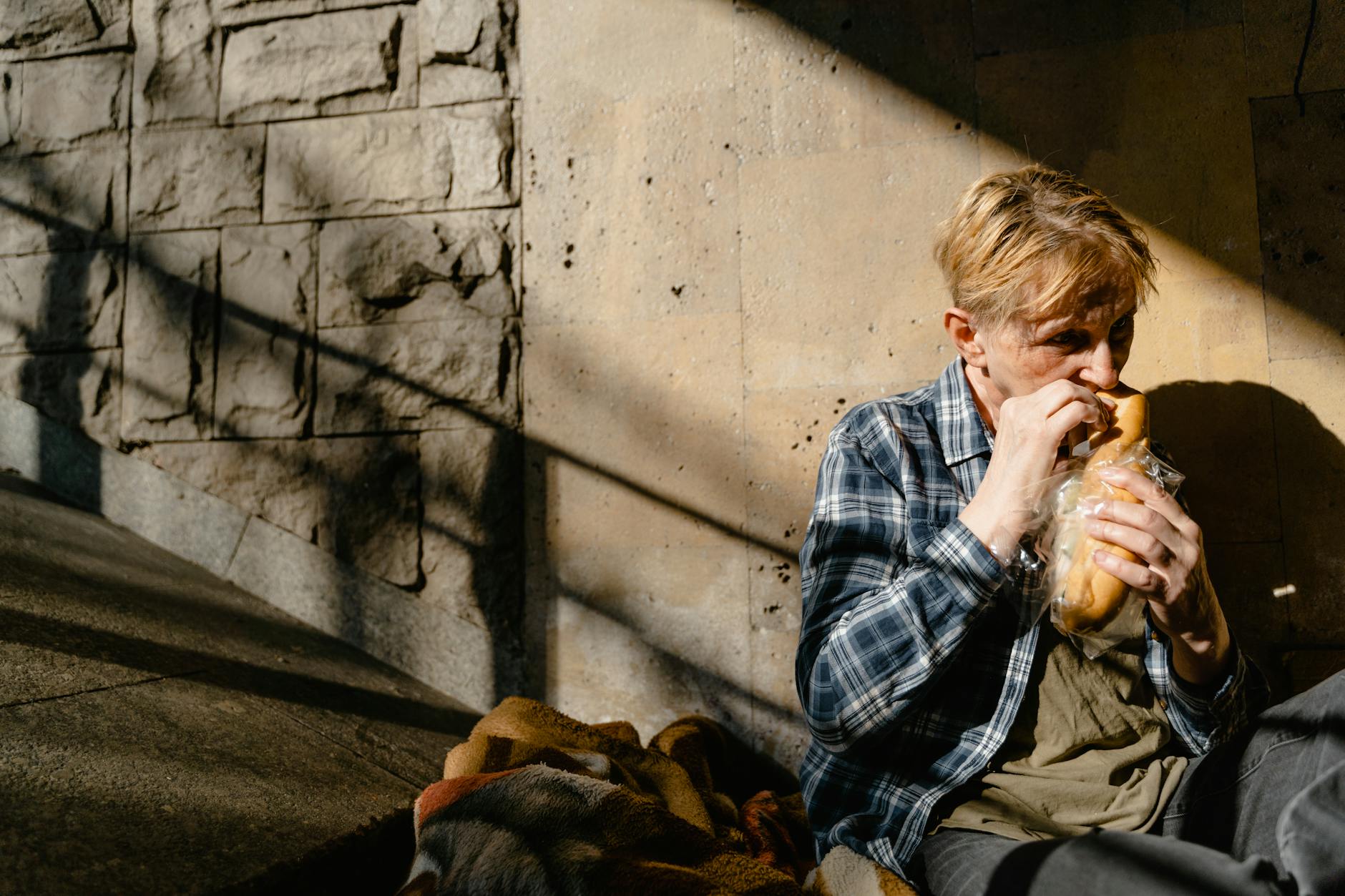 fragilità sociale italiana — A senior man sitting against a wall, eating bread, highlighting homelessness.