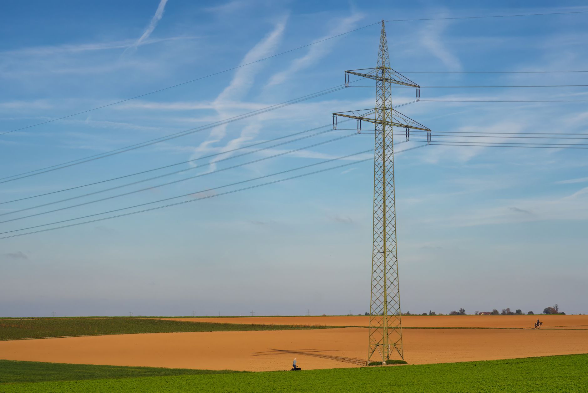 deroghe patto stabilità — High voltage power line standing tall in a vast agricultural field under a clear blue sky in Germany.