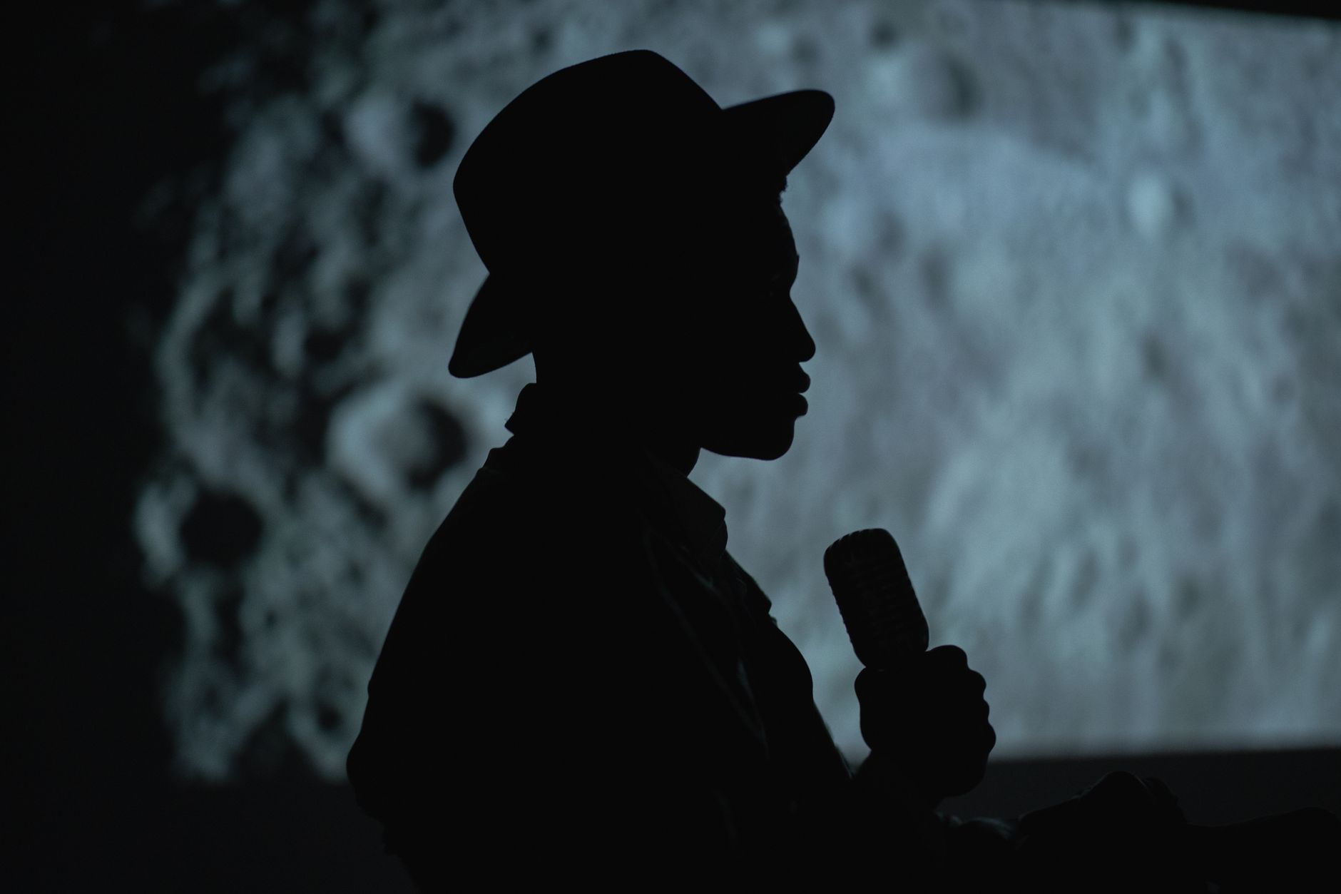 performance poetica — A dramatic silhouette of a person with a hat holding a microphone against a moon-like background.