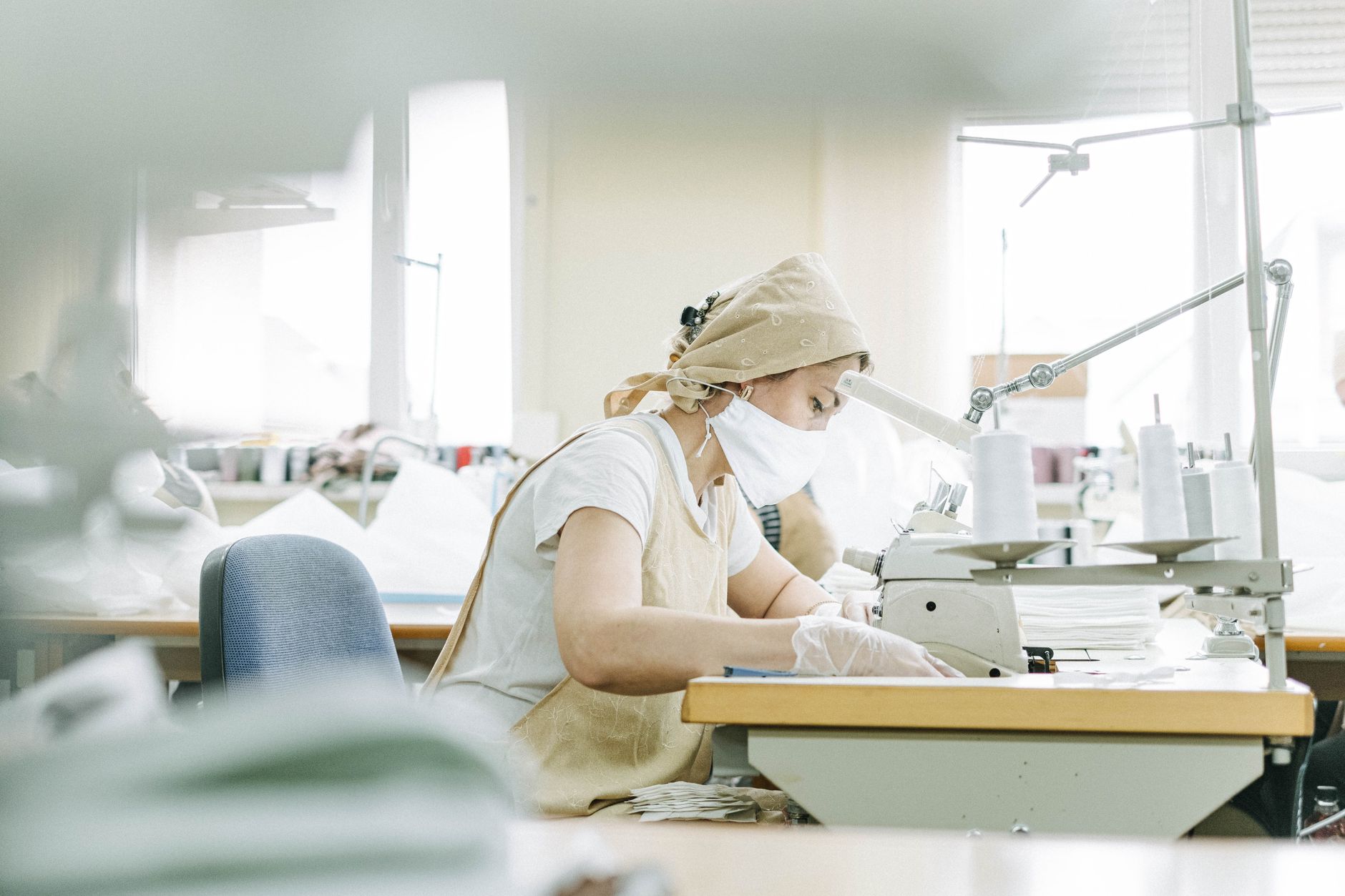 sistema moda italiano — A focused seamstress wearing protective gear works diligently at a sewing machine in a factory setting.