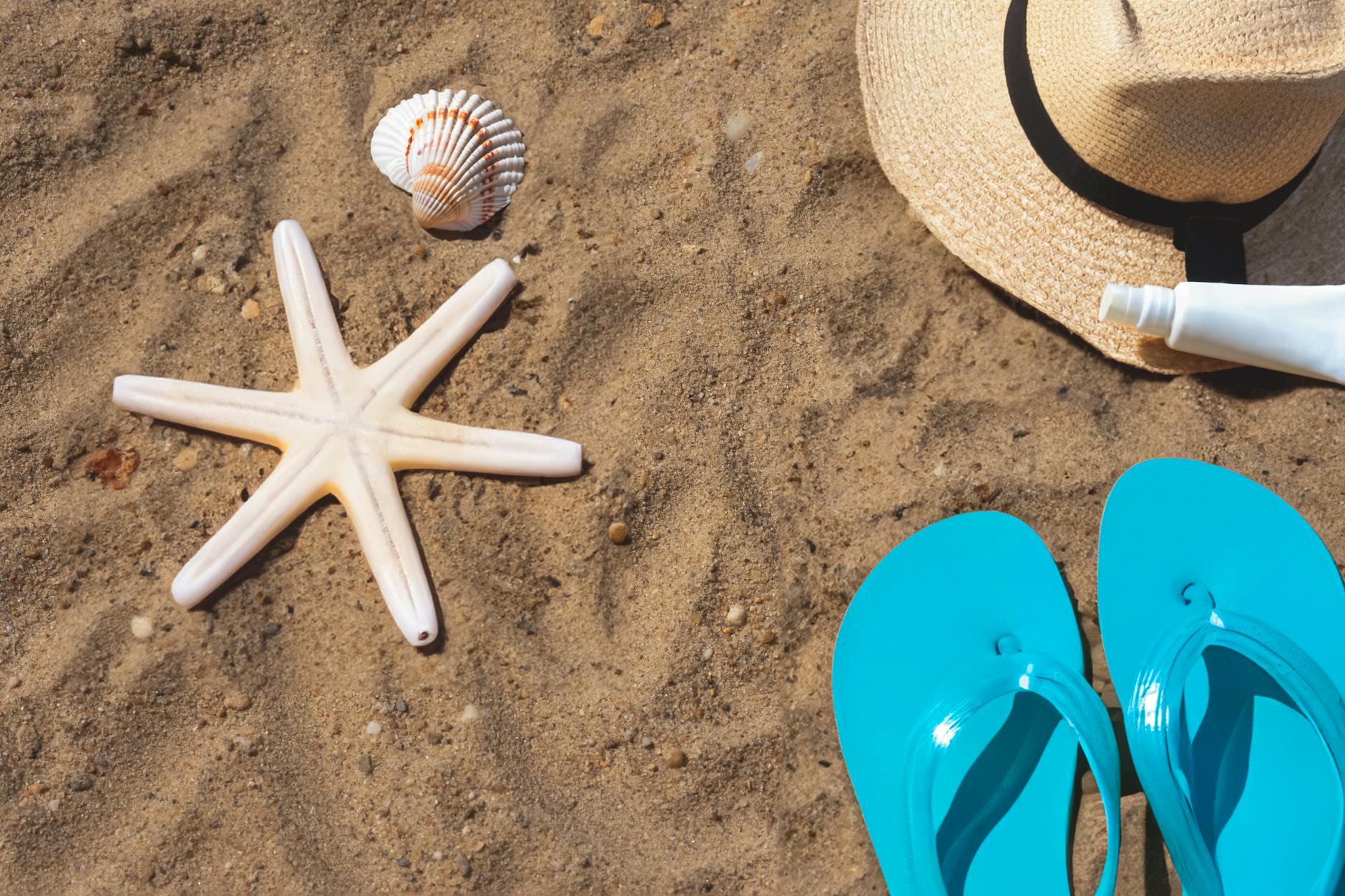 gadget indispensabili viaggio — Flat lay of beach essentials including flip flops, hat, starfish, and shells on sand.