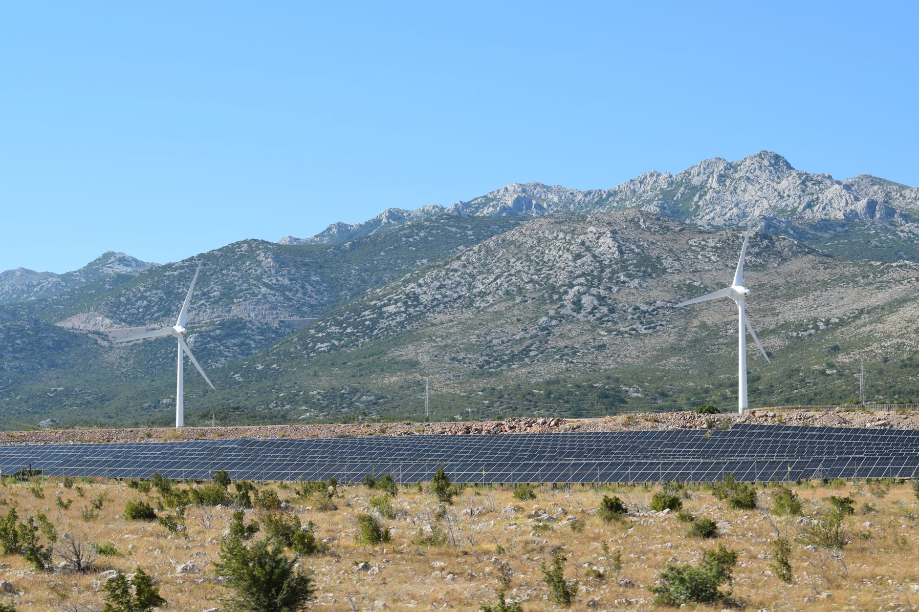investimenti rinnovabili — A scenic view of windmills and solar panels against mountainous terrain under a clear blue sky.
