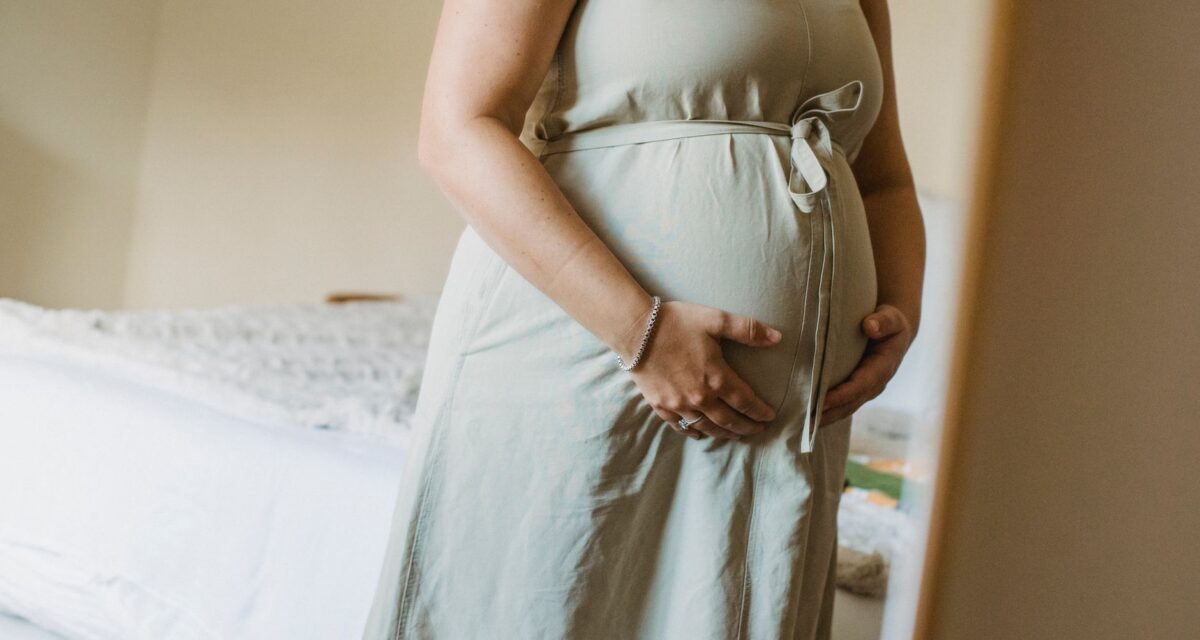 Faceless crop adult pregnant female standing near mirror in room while touching stomach in daytime